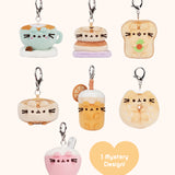 Front view of Pusheen Breakfast Surprise Plush assortment. Pusheen the Cat has taken the form of seven miniature breakfast-inspired plush varieties. From left to right and top to bottom, the surprise plush are a Latte, Pancake Stack, Avocado Toast, Waffle, Orange Juice, Sesame Bagel, and Bowl of CerealThe bottom right corner has a heart with the words “1 Mystery Design!” indicating eight possible miniature plush keychains are available.