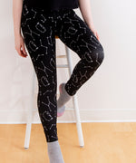 Model wearing the Pusheen Celestial Leggings. The black leggings feature a pattern of various white star constellations and white line art of Pusheen hopping in different directions. The model is sitting on a stool in front of a white background.