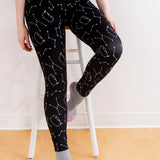 Model wearing the Pusheen Celestial Leggings. The black leggings feature a pattern of various white star constellations and white line art of Pusheen hopping in different directions. The model is sitting on a stool in front of a white background.