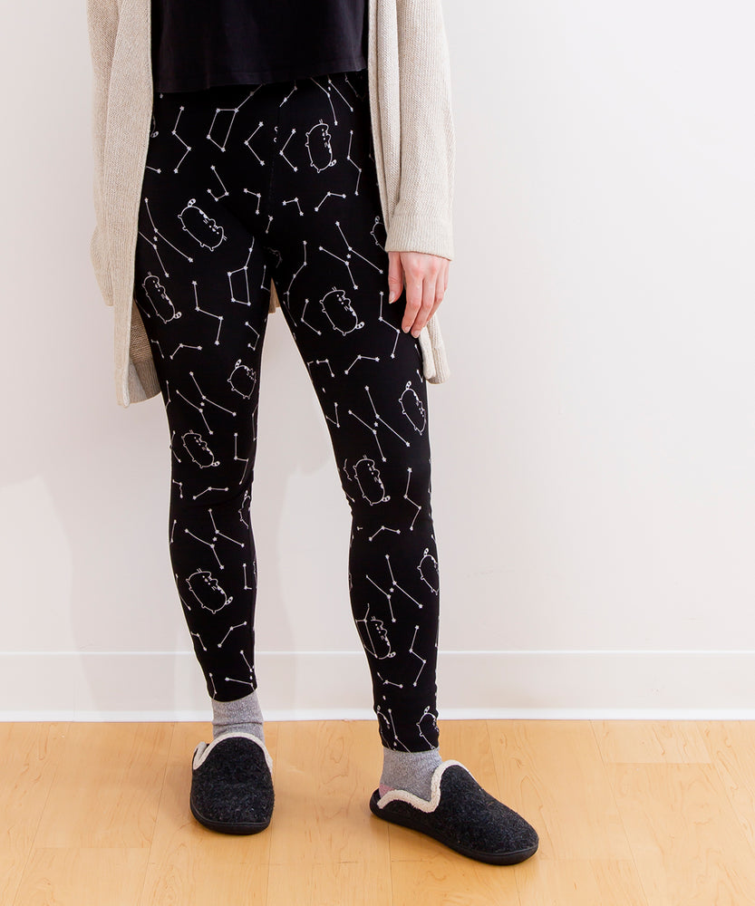 Model standing while wearing the Pusheen Celestial Leggings. The model is wearing a loose knit sweater and black slippers with a white trim. The model is standing in front of a white wall and a wooden floor.