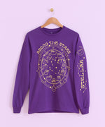 The Pusheen Celestial Long-Sleeve Unisex Tee hangs on a brown hanger in front of a light purple wall.  The dark purple tee shirt features Pusheen the Cat as the zodiac signs in an astrological chart. The large front graphic is gold and is accompanied by a gold graphic down the wearer’s left sleeve. 