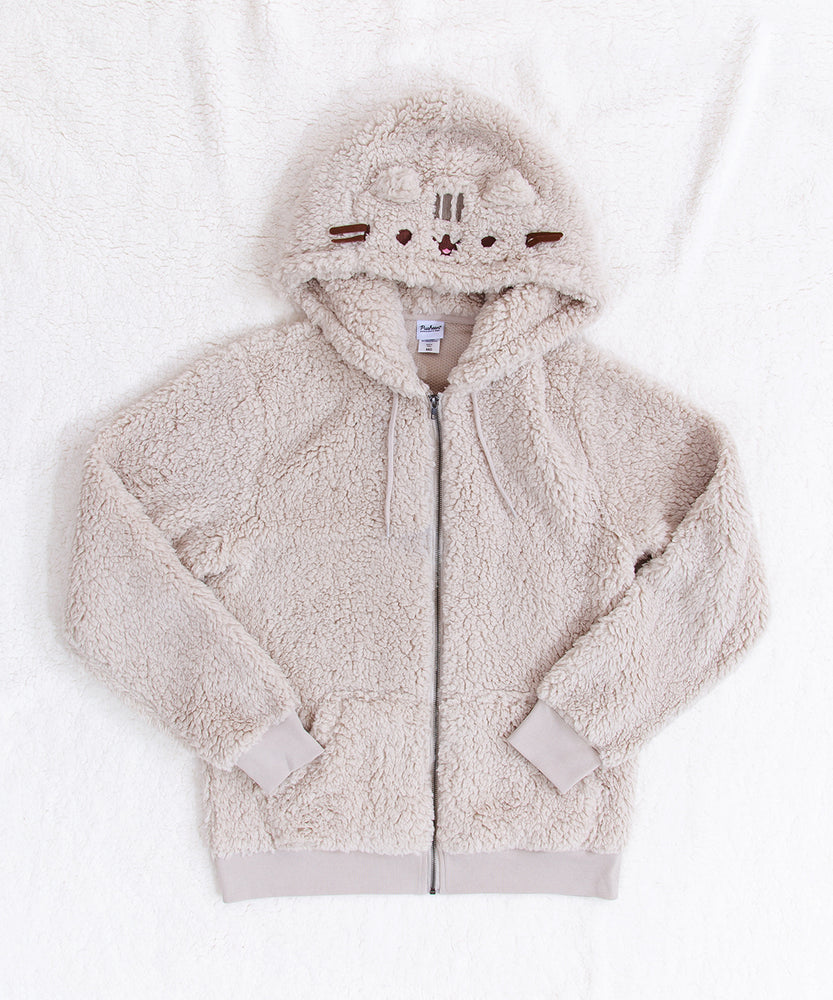 A fuzzy light grey zip up hoodie with cat ears and Pusheen's face on the hood. There are drawstrings on the hoodie, pockets, and the cuff and waistband are ribbed fabric. The hoodie is laying on top of a fuzzy white blanket with the sleeves folded inwards.