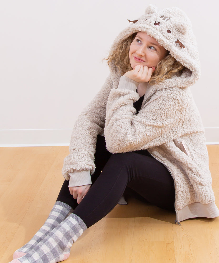 Model sitting on a wooden floor, wearing the Pusheen Character Hoodie with the hood pulled up. The model is smiling while daydreaming. 