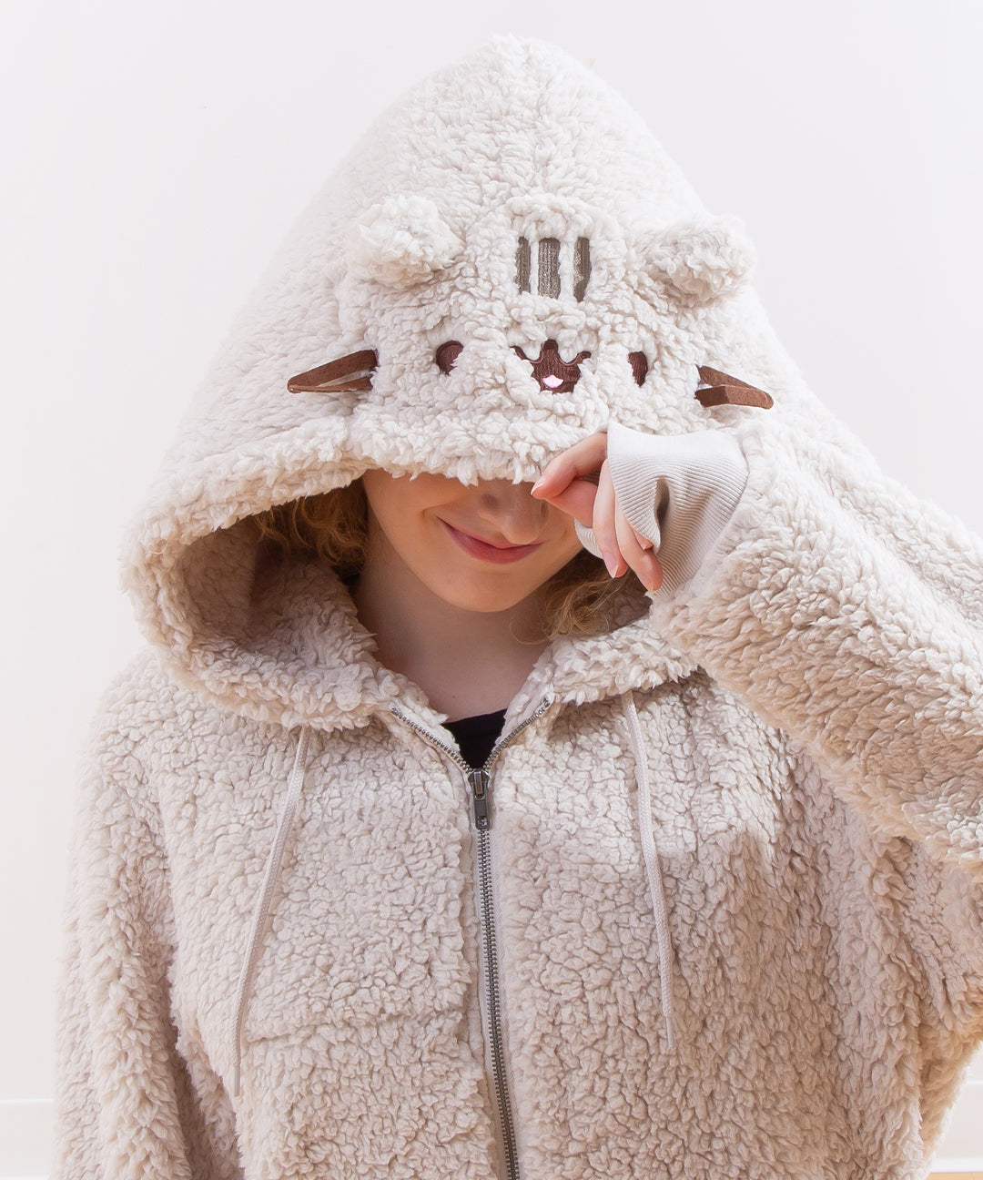 Close up of a model wearing the Pusheen Character Hoodie, smiling as they pull down the hood over their eyes. Pusheen's features are closer to the edge of the hood.