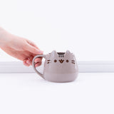 A hand holding the Pusheen mug’s handle from the left in front of a white background. The mug takes on more of a trapezoid shape, with a wider base and a more narrow opening. The mug is very shiny and reflects light.
