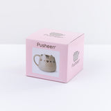 The box packaging for the Pusheen Character Mug in front of a white background. The box is a pink cardboard square that cover the mug entirely. On the front of the box is the Pusheen logo, a photo of the mug inside, and the name ‘Ceramic Mug’ printed underneath it in English and French. The top and side of the box features the Pusheen the Cat Logo.