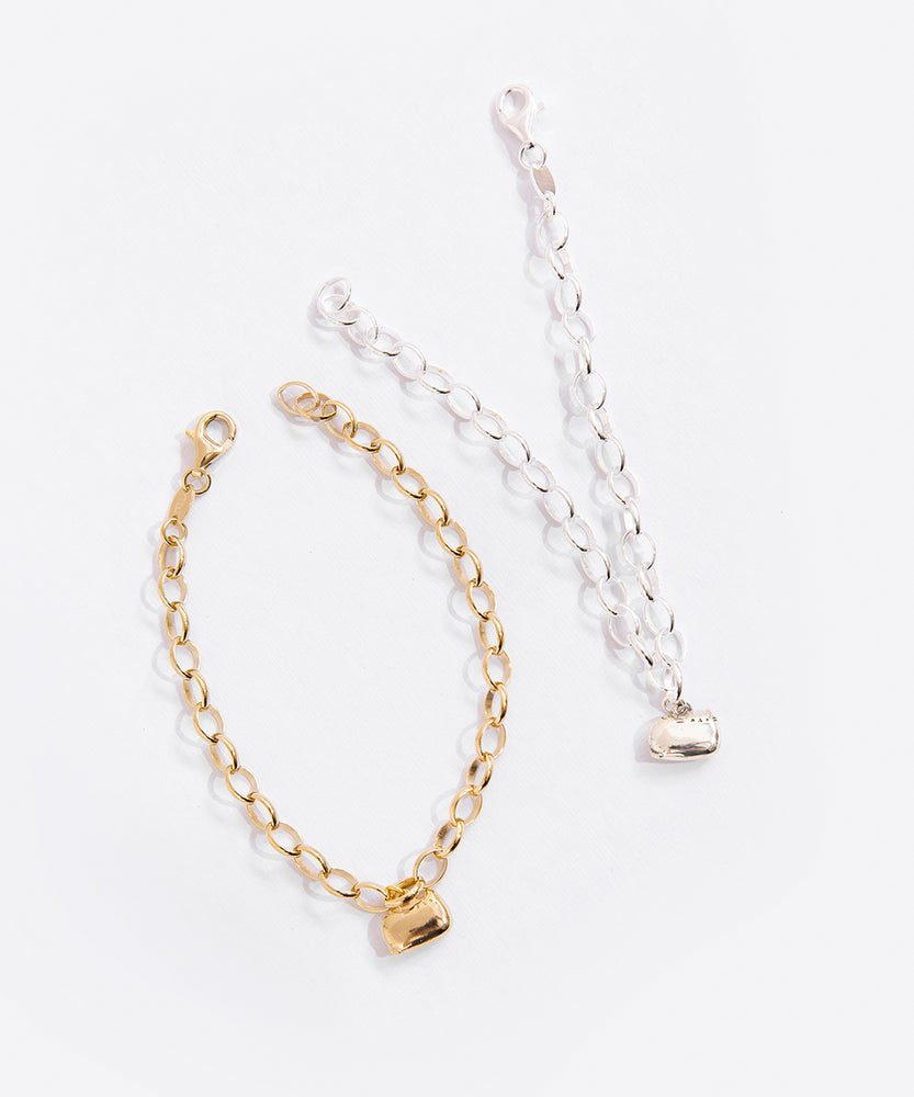 Gold and Silver charm bracelets next to one another, with the gold chain curved like a circle and the silver chain pointed out in a 'v' shape. Both chains use a lobster claw clasp attached to an oval shape.