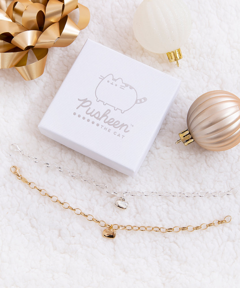 The gold and silver charm bracelets unclasped besides one another and underneath the square white jewelry box featuring the Pusheen the Cat logo printed in silver.  The items are on top of a plush white blanket, and surrounded by white and bronze Christmas ornaments and a golden gift bow.