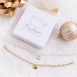 The gold and silver charm bracelets unclasped besides one another and underneath the square white jewelry box featuring the Pusheen the Cat logo printed in silver.  The items are on top of a plush white blanket, and surrounded by white and bronze Christmas ornaments and a golden gift bow.