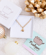 The Gold Charm Bracelet in a white square jewelry box. The jewelry lid is partially on top of the box, and the Pusheen the Cat cart insert for the jewelry box is underneath the bottom right of the box.The box is surrounded by a shiny gold gift bow and white and bronze holiday ornaments, all on top of a plush white blanket.