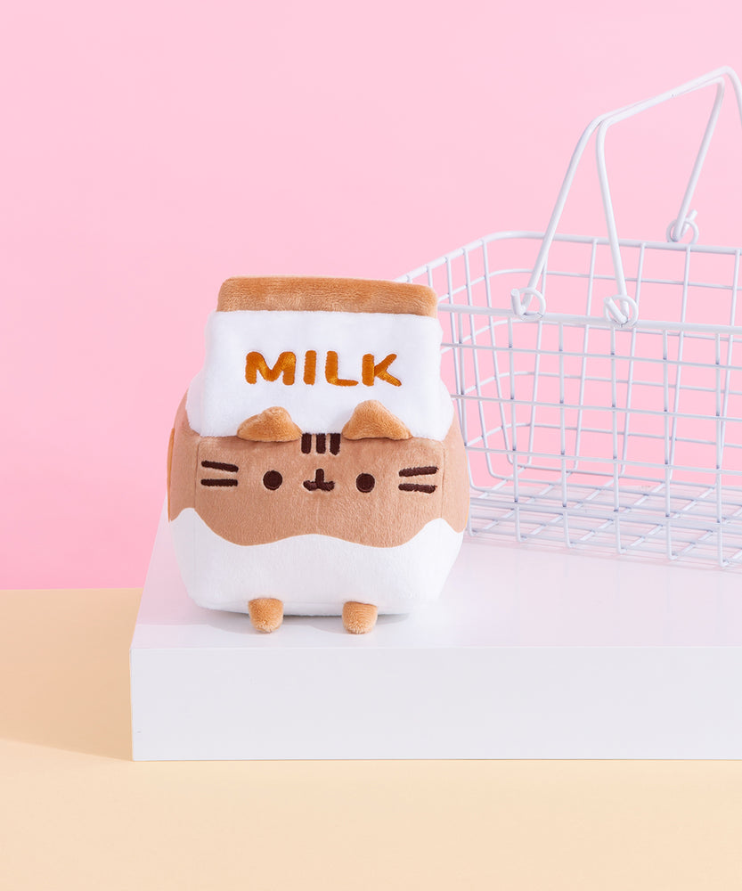 The Pusheen Chocolate Milk Sips Plush seated on a white pedestal with a small white wire basket in the background. The plush is in the shape of a small milk carton with a brown and white wave throughout the body and the word ‘Milk’ embroidered in brown stitching near the carton lid. Pusheen’s face is embroidered in dark brown on the plush, with plush ears at the rim of the carton lid and brown plush feet at the bottom. 