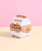 Sitting in front of a pink and yellow background, Chocolate Milk Sips Plush is showing its side profile that showcases an embroidered chocolate square detail.  