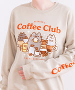 Model wearing the Pusheen Coffee Club Unisex Sweatshirt. The tan sweatshirt features a large graphic in the center of the sweatshirt of Pusheen the Cat, Little Brother Pip, and Little Stormy enjoying coffees and sweet treats. The phrase “Pusheen’s Coffee Club” sits above the cats in red orange text while the phrase “Catfé Est. 2010” sits below the graphic. The model is holding their arm up to show a printed sleeve detail featuring the phrase “Coffee O’Clock”. 