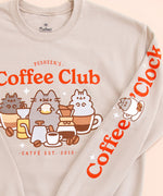 Close-up view of the front and sleeve graphics of the Pusheen Coffee Club Unisex Sweatshirt. The grey cats are surrounded by coffee making machines, drinks, cups, and coffee beans. Pusheen eats a yellow and white cinnamon roll.  