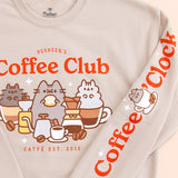 Close-up view of the front and sleeve graphics of the Pusheen Coffee Club Unisex Sweatshirt. The grey cats are surrounded by coffee making machines, drinks, cups, and coffee beans. Pusheen eats a yellow and white cinnamon roll.  