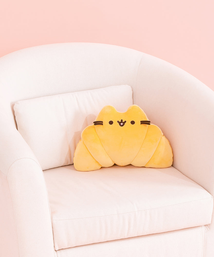 Croissant Pusheen Squisheen sits on a cream chair. The yellow croissant-shaped plush has brown embroidery features for Pusheen's eyes, mouth, and whiskers.