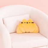 Croissant Pusheen Squisheen sits on a cream chair. The yellow croissant-shaped plush has brown embroidery features for Pusheen's eyes, mouth, and whiskers.