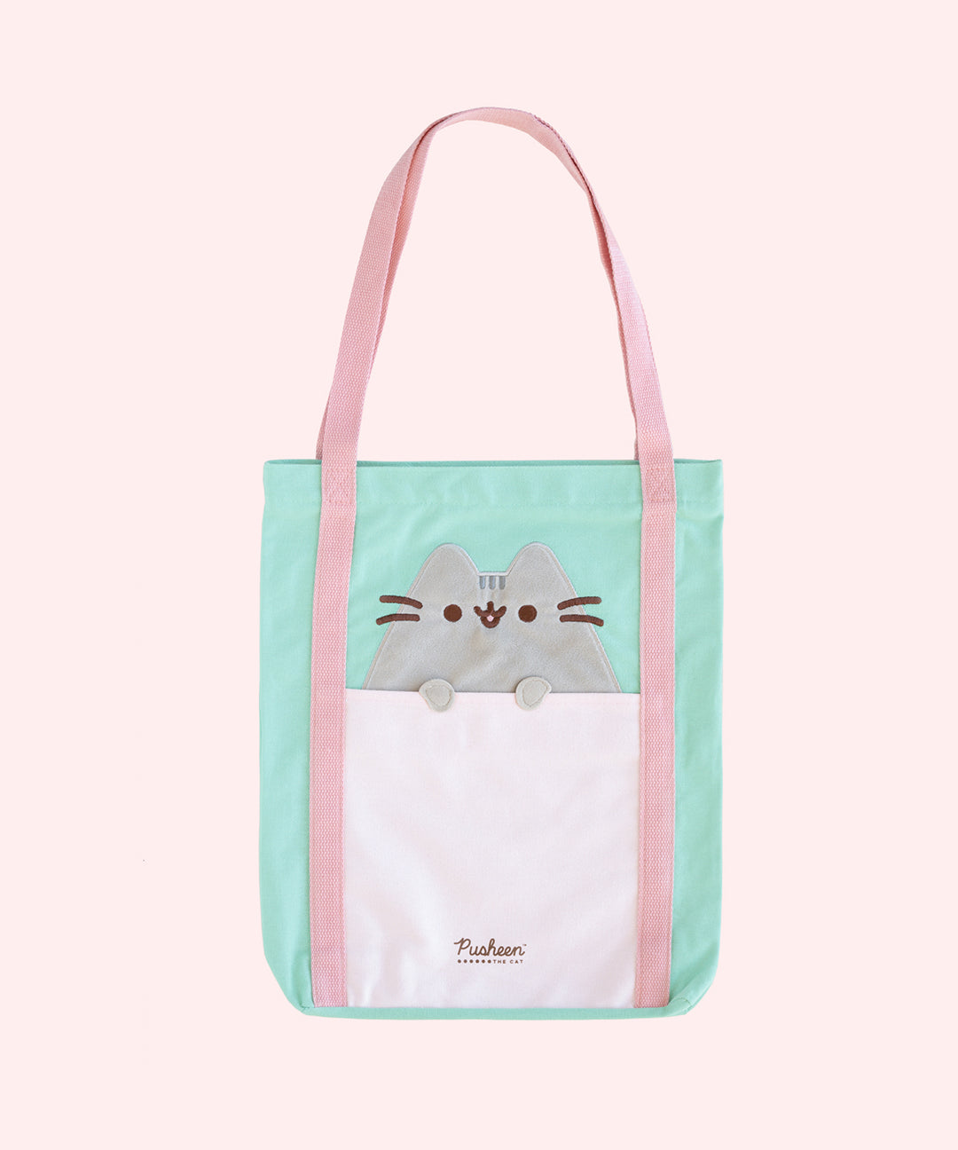 A tote bag is propped up on a pink surface. The mint green tote has an applique of Pusheen the Cat with her paws hanging over the front pocket. The handles of the bag are pink, and the base has the Pusheen the Cat logo in brown. 