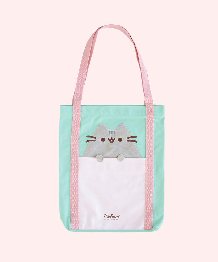 A tote bag is propped up on a pink surface. The mint green tote has an applique of Pusheen the Cat with her paws hanging over the front pocket. The handles of the bag are pink, and the base has the Pusheen the Cat logo in brown. 