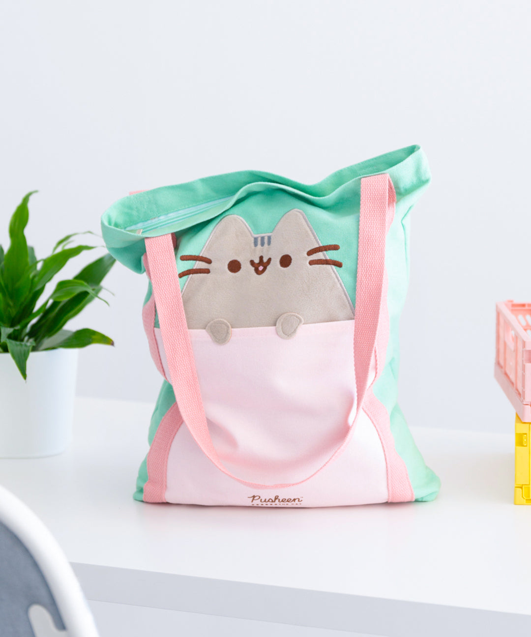 Front view of the Pusheen Deluxe Tote Bag. The mint green tote bag has a light pink front pocket and pink handles that extend down the length of the bag. A grey Pusheen the Cat applique sticks out of the front pocket with her paws hanging over the opening. 