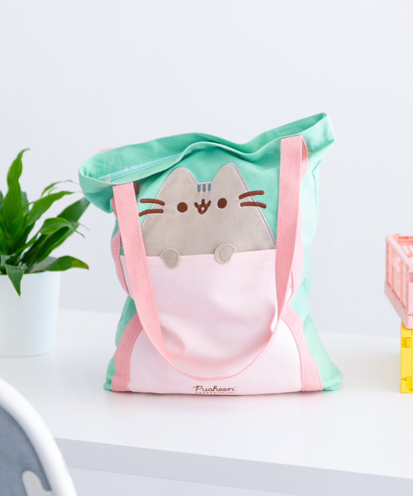 Front view of the Pusheen Deluxe Tote Bag. The mint green tote bag has a light pink front pocket and pink handles that extend down the length of the bag. A grey Pusheen the Cat applique sticks out of the front pocket with her paws hanging over the opening. 