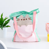 Front view of the Pusheen Deluxe Tote Bag. The mint green tote bag has a light pink front pocket and pink handles that extend down the length of the bag. A grey Pusheen the Cat applique sticks out of the front pocket with her paws hanging over the opening. 