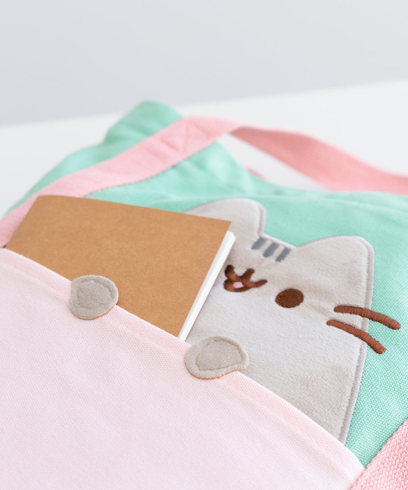 A close-up of the light pink front pocket of the tote. Pusheen's face is shown behind the pocket. A brown notebook is sticking out of the front pocket to show the paws applique. 