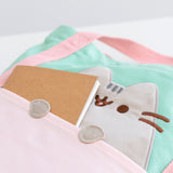 A close-up of the light pink front pocket of the tote. Pusheen's face is shown behind the pocket. A brown notebook is sticking out of the front pocket to show the paws applique. 