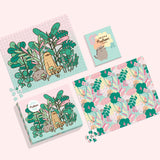 The Pusheen Double-Sided 500-Piece Puzzle box, sticker book, and both sides of the puzzle lie on a light pink surface. All elements feature graphic of Pusheen surrounded by plants and pink, yelllow, orange, and light blue details.  