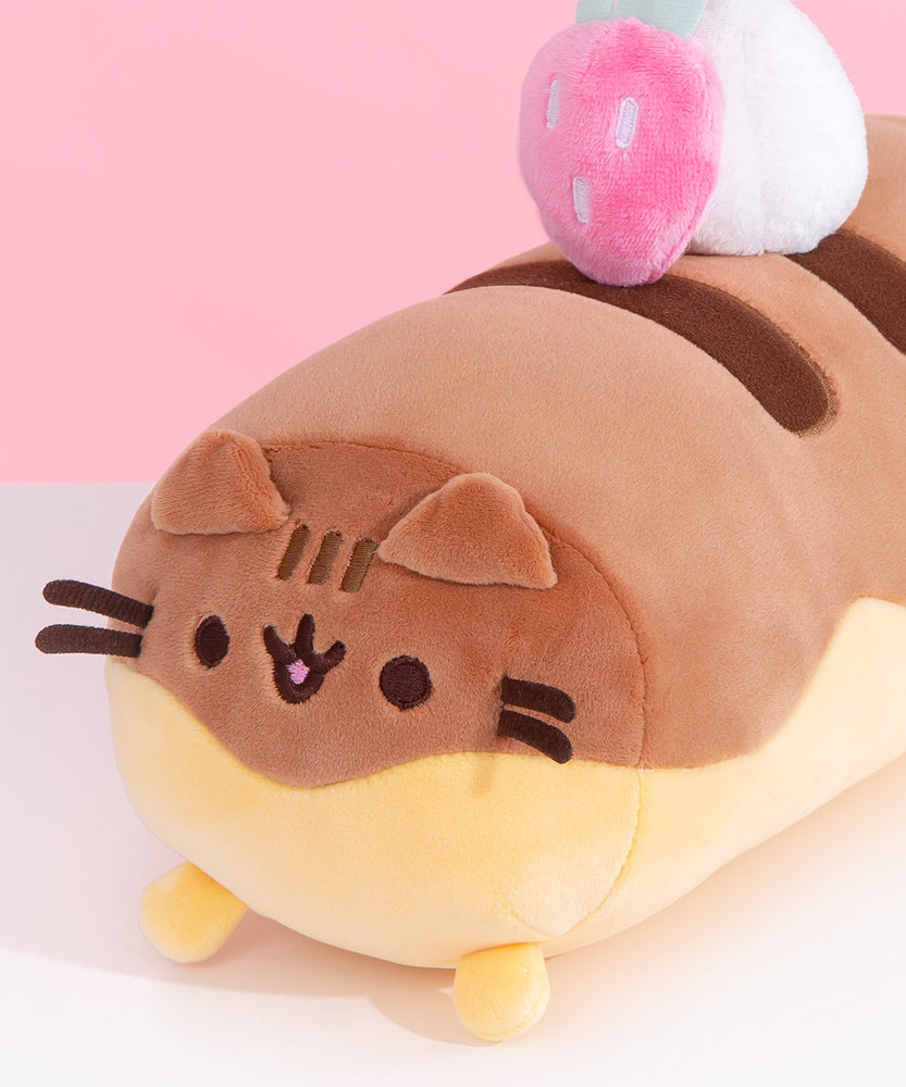 Close-up view of Pusheen Éclair Squisheen plush. Pusheen takes the form of a delicious dessert and her classic ears, eyes, stripes, mouth, and whiskers are embroidered at the end of the desert plush.