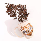Pusheen Espresso Yourself Mug lies on a white surface with coffee beans coming out of the top. The tan mug features a printed graphic Pusheen, Little Brother Pip, and Little Sister Stormy as coffee drinks. The trio is accompanied by brown coffee beans and the phrase “Espresso Yourself” in brown text. 