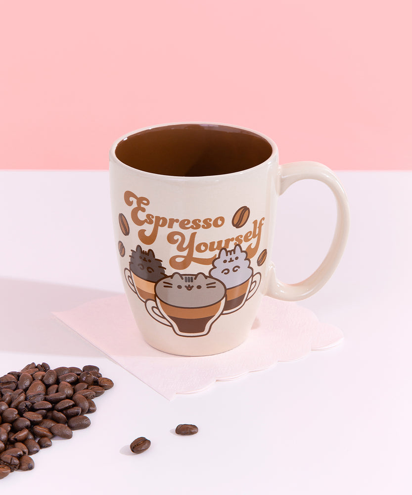 Front view of the tan Pusheen coffee mug. The interior of the mug is a darker brown color compared to the light tan exterior of the mug. The graphic of Pusheen, Pip, and Stormy covers the entire front side of the mug and ends near the handle. 