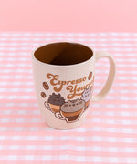 Top view of the Pusheen mug. In the printed graphic, the trio of cats sit inside various coffee beverages that are half coffee and half additional liquids. The tan and brown cup sits atop a pink and white checkered surface in front of a pink background.  