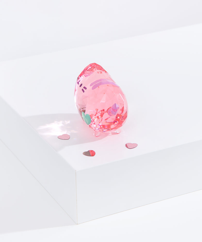 Side view of Pusheen Facets shows the width of the figurine scale. Figurine standing on a white surface surrounded by pink confetti hearts. Facets geometric reflection can be seen on white surface. 