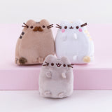 Close up of the mini plush, Pusheen’s parents on top of the pink pedestal while Pusheen sits underneath them on the white floor. The parent’s poses mirror each other, with the father’s tail pointing out from the left and the mother’s tail pointing out from the right. Pusheen faces straight forward, no tail visible and mouth open.