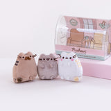 The three mini plush outside of the packaging , standing next to one another in front of a white background, with the original packaging still intact and on top the pink pedestal. Pusheen’s father has his feet on the ground, while Pusheen and her mother have all four paws facing upfront. The illustrated background in the packaging also features two arm chairs and a mini table with two tea mugs on top of it.