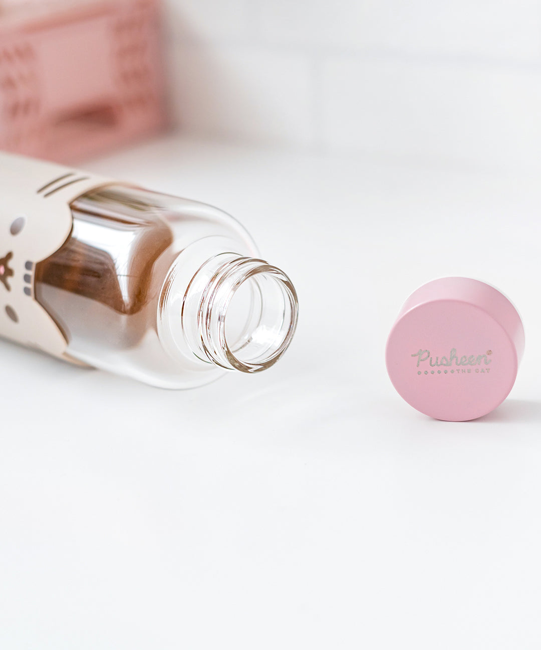 Glass water bottle lies on a white surface showing the inside of the open bottle. The pink top features a Pusheen the Cat logo in silver print.  