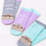 Close-up view of all three sock sets from the Pusheen Good Vibes 3-Pack Ankle Socks that are laid flat in pairs on a white fluffy surface. From left to right are the grey and purple “good vibes” ankle socks, the “let’s stay in” purple and light green sock set, and the light green and grey Pusheen spectacles glasses.  