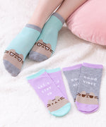 A model wears a pair of ankle socks besides two pairs of ankle socks, all on top of a fluffy white carpet. The model is wearing light green socks with grey accents with a graphic of Pusheen wearing glasses. One pair of socks by the model are purple with light green accents. This sock set features the phrase “let’s stay in” above a Pusheen graphic. The last pair of socks is grey with purple details. This sock set features Pusheen wearing sunglasses paired with the phrase “Good Vibes." 