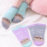 A model wears a pair of ankle socks besides two pairs of ankle socks, all on top of a fluffy white carpet. The model is wearing light green socks with grey accents with a graphic of Pusheen wearing glasses. One pair of socks by the model are purple with light green accents. This sock set features the phrase “let’s stay in” above a Pusheen graphic. The last pair of socks is grey with purple details. This sock set features Pusheen wearing sunglasses paired with the phrase “Good Vibes." 