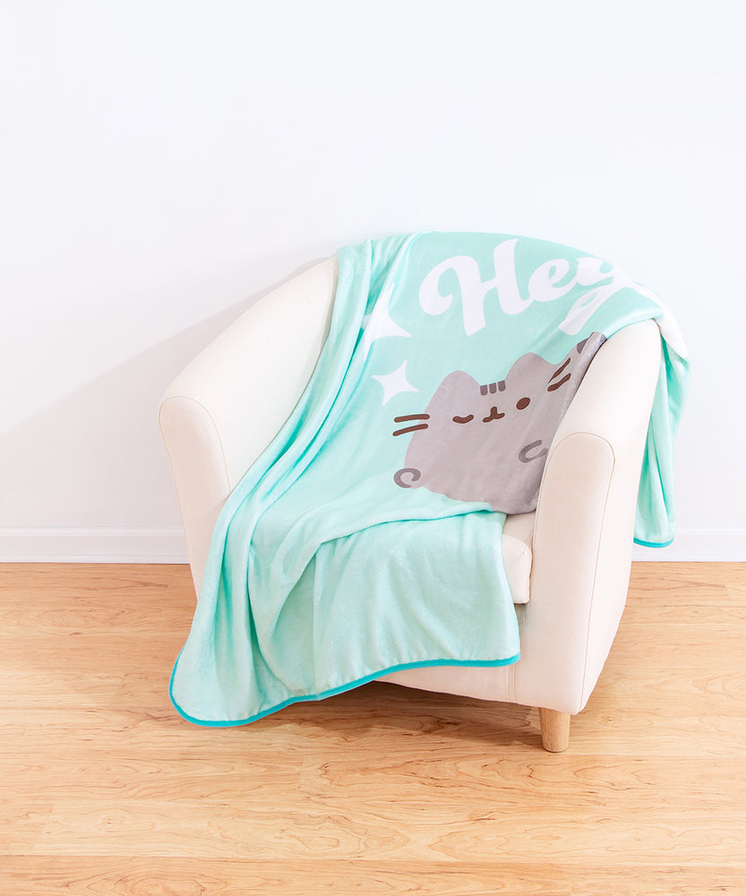 Pusheen Hey Throw Blanket draped on top of a cream chair. The teal blanket features a graphic of Pusheen the Cat winking while saying “Hey.” The center graphic is surrounded by white stars.  