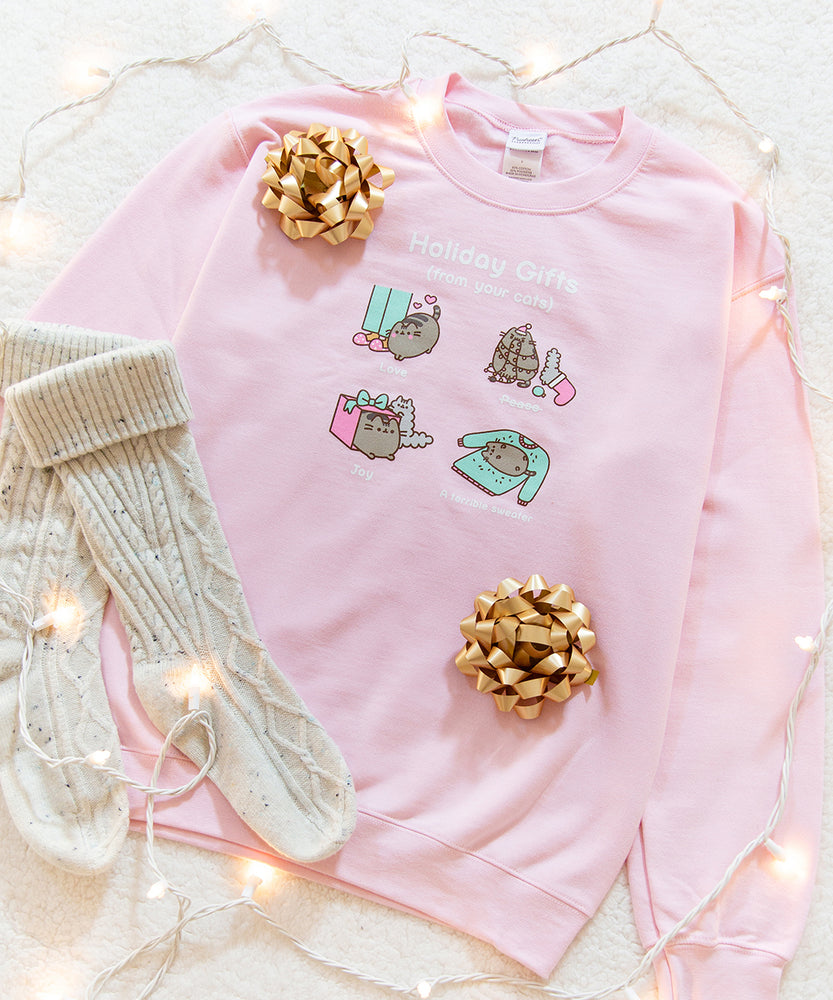Front view of the pink unisex sweatshirt is surrounded by cozy knit socks, twinkling lights, and hold present bows. The peace gift from Pusheen shows the cat tangled up in Christmas lights while Stormy rummages through a holiday stocking.  