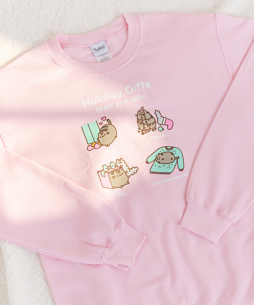 Close-up view of the holiday gifts graphic in the front center of the pink sweatshirt. The terrible sweater gift shows Pusheen rubbing against a person’s sweater leaving a mess of her hair behind. Sunlight shines on the graphic of the sweatshirt.  
