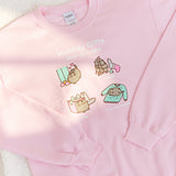 Close-up view of the holiday gifts graphic in the front center of the pink sweatshirt. The terrible sweater gift shows Pusheen rubbing against a person’s sweater leaving a mess of her hair behind. Sunlight shines on the graphic of the sweatshirt.  
