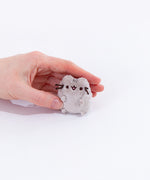 A model’s hand gently covering the mini Pusheen plush from the collectors set. The plush is no bigger then the top of the fingers. Pusheen’s face and head stripes are embroidered into the mini plush, and her whiskers are made of stiff string. 