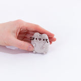 A model’s hand gently covering the mini Pusheen plush from the collectors set. The plush is no bigger then the top of the fingers. Pusheen’s face and head stripes are embroidered into the mini plush, and her whiskers are made of stiff string. 