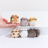 The I Love Kitties mini plush lined up in two rows of 3, a model’s hand holding the winking brown mini cat plush on the tip of their fingers. All of the mini plush have all of their paws sticking out forward, save for the annoyed white cat who has one paw over their mouth and nose.