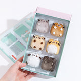 A model’s hand holding the mini plush in their original container with the lid removed. The container is a plastic tray with three rows of two within a pink and mint cardboard box, with the mini plush resting inside like ice cubes in a tray. The top row features Pusheen and the winking brown cat, the middle row has the Siamese cat and the worried yellow cat, and the bottom row has the annoyed white cat and the skunk.