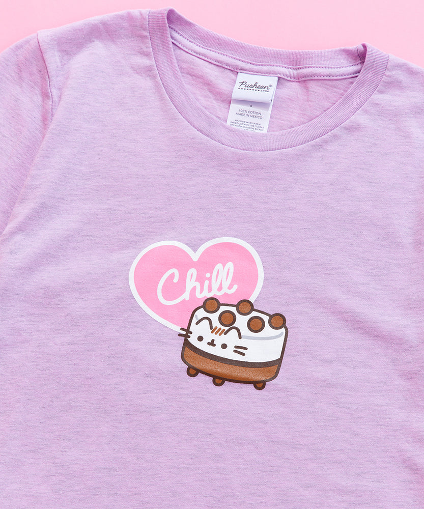 A close-up view of the tee’s screen print graphic. Ice Cream Cake Pusheen is a short cylinder shape with white and brown striped details with light brown balls atop the white top. The ice cream cake features Pusheens’ face printed on the front, her striped tail sticking out from the back, her paws at the bottom of the cake. The word ‘Chill’ is printed in white in a light pink heart with a white outline. The pink heart accompanies the ice cream cake in the center of the tee shirt. 
