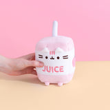 A model’s hand lightly squeezes the Juice Box Sips Plush in front of a pink and yellow background.  Pusheen is in the shape of a juice box, with a plush straw sticking out on top. The top and bottom of the box are light pink, and the middle is white, with an embroidery of Pusheen’s face, the word ‘juice’ in pink, and a partially visible graphic on the right side. There are white plush ears sticking out under the rim of the box, and pink plush feet sticking out from the bottom. 
